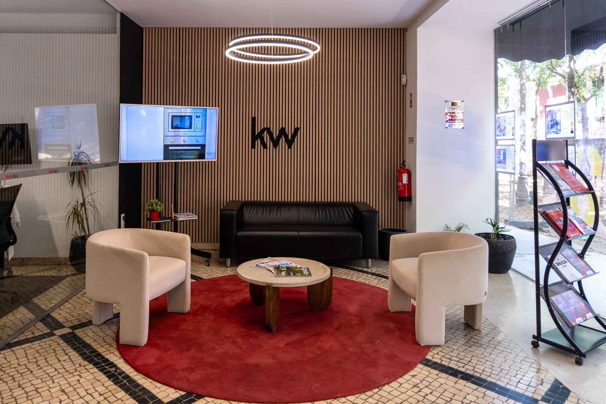 Sala de espera do piso 0 da KW Area: duas poltronas claras e uma mesa redonda sobre um tapete encarnado, com o símbolo kw na parede de ripas de madeira